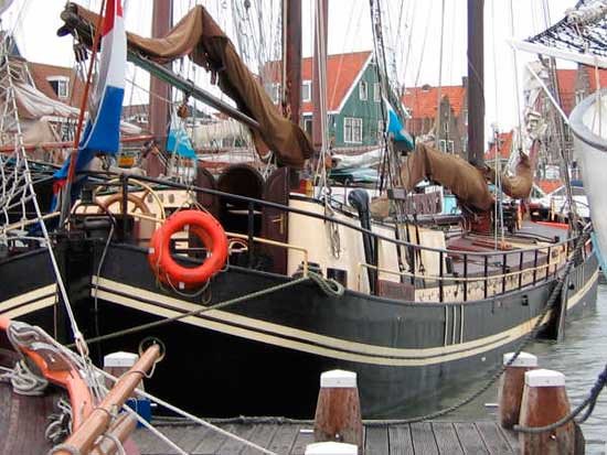 The ship moored in a small harbour with green wooden houses behind, an orange life ring on the rail