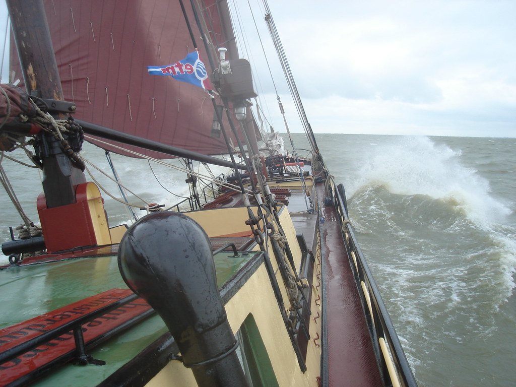 The bow of De Chateauroux cutting through a wave under red sails